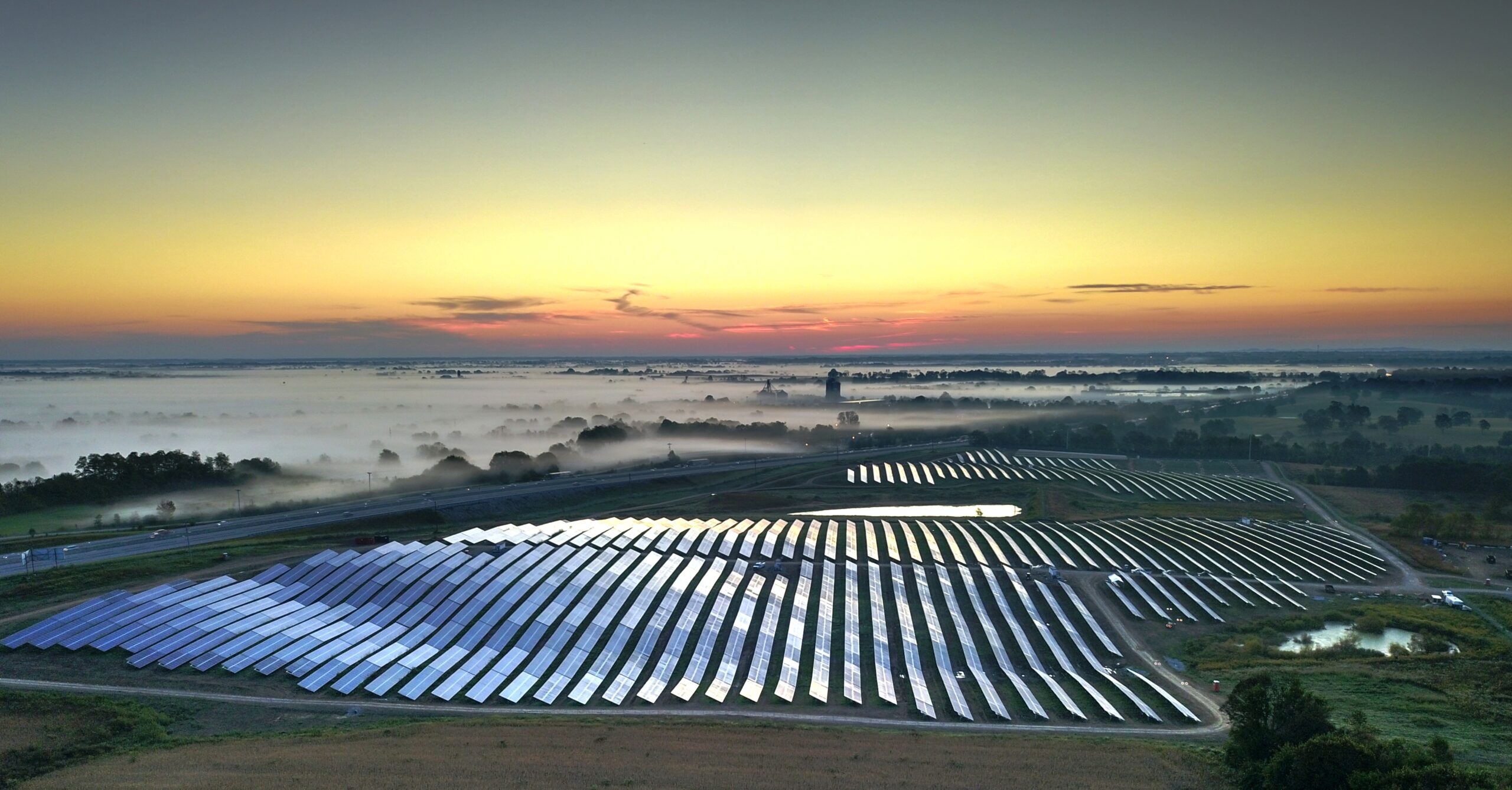 The sun rises over a solar farm, casting a warm glow on the rows of solar panels as misty fog blankets the surrounding landscape.